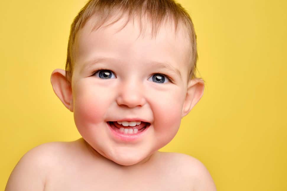 Smiling baby model with blue eyes and baby teeth in a close-up portrait against a bright yellow background.