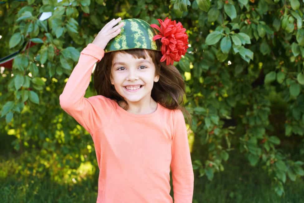 Smiling child model wearing a watermelon hat with a red flower, posing outdoors in a playful and cheerful modelling photo.