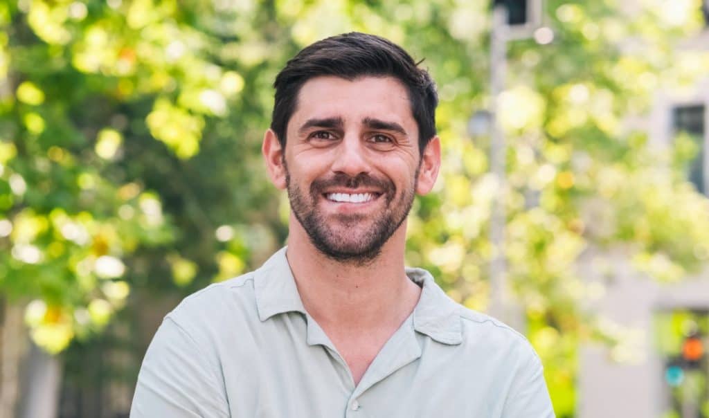 miling male model with a beard in a casual shirt, posing outdoors in natural light with a blurred green background.