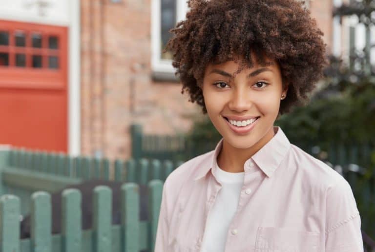 Confident young woman with curly hair smiling outdoors in a casual shirt, posing for a lifestyle modelling headshot.